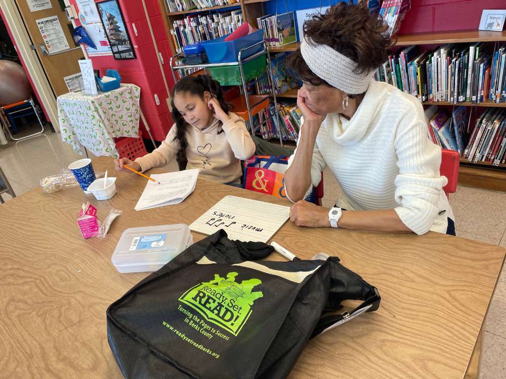 An adult volunteer and a young student work together at a wooden table as part of the Ready.Set.READ! program. A black tote bag with the program's logo sits in the foreground, while the background shows colorful library bookshelves. The student is focused on a worksheet while the tutor observes, with a small whiteboard nearby showing the words "music notes."