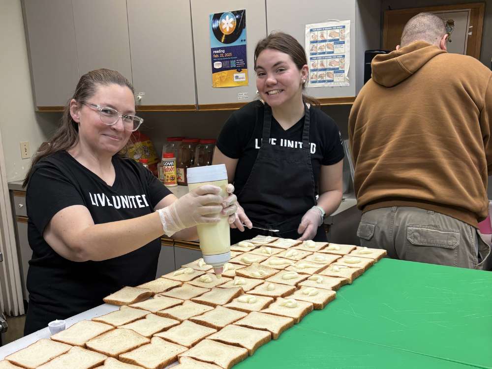 Three United Way of Berks County volunteers wearing bright orange safety vests smile while holding donated food items in a large warehouse. The group includes two women and a man, all holding various yellow food boxes and jars. In the background, tall industrial shelves are stacked high with pallets of canned goods and supplies.