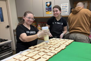 Two Live United volunteers wearing black t-shirts smile while preparing food in a kitchen. One volunteer uses a large squeeze bottle to apply a cream topping to dozens of slices of bread neatly arranged on a counter, while the other stands beside her.