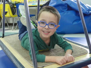 A smiling young boy in bright blue glasses and a green shirt lies on a small rug inside a classroom play structure.