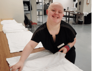 A smiling young woman folding white linens on a long wooden table.