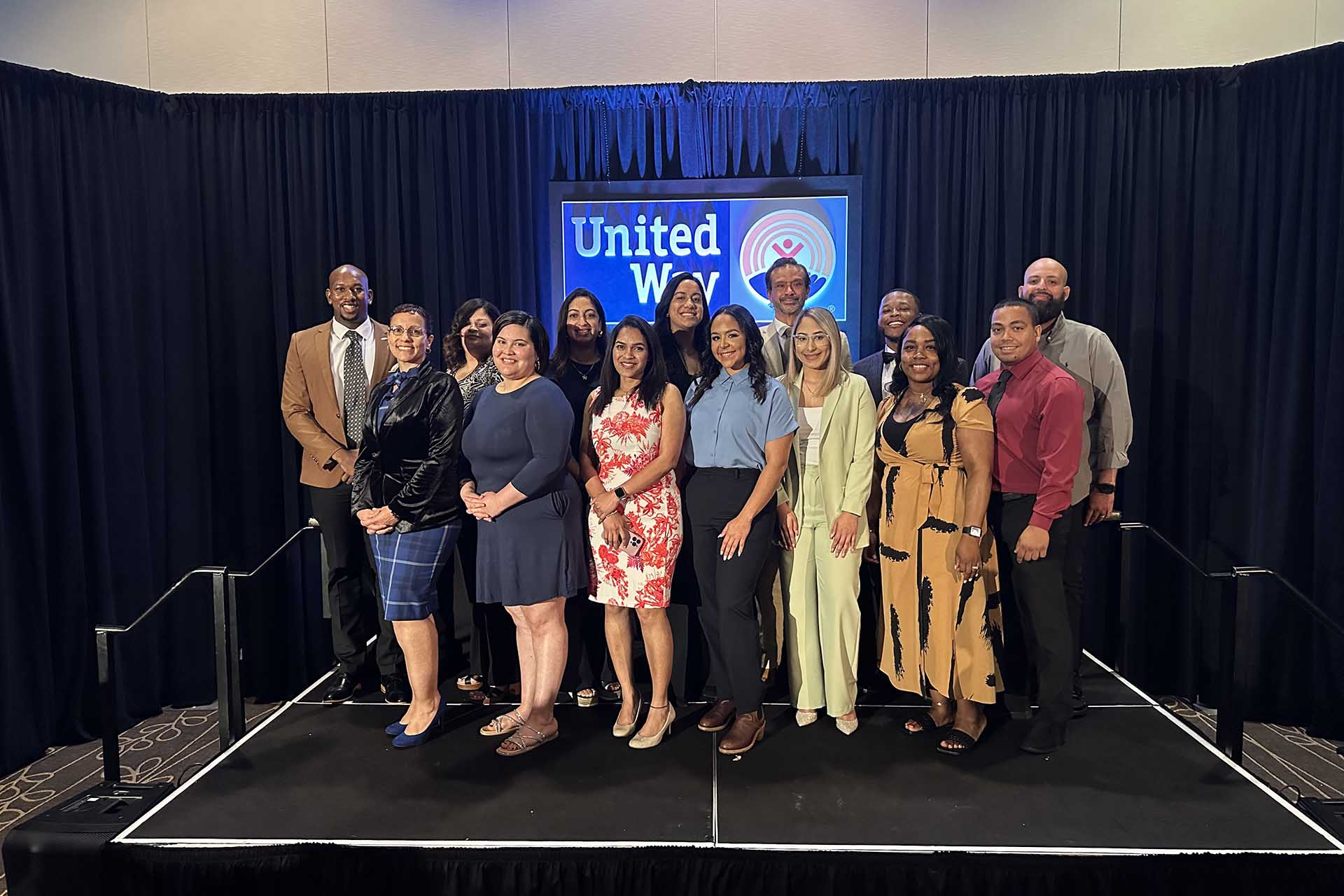 A diverse group of about 14 professionally dressed individuals posing for a group photo on a small stage. They are standing in front of black curtains, and a digital screen behind them displays the United Way logo.</p>
<p>
