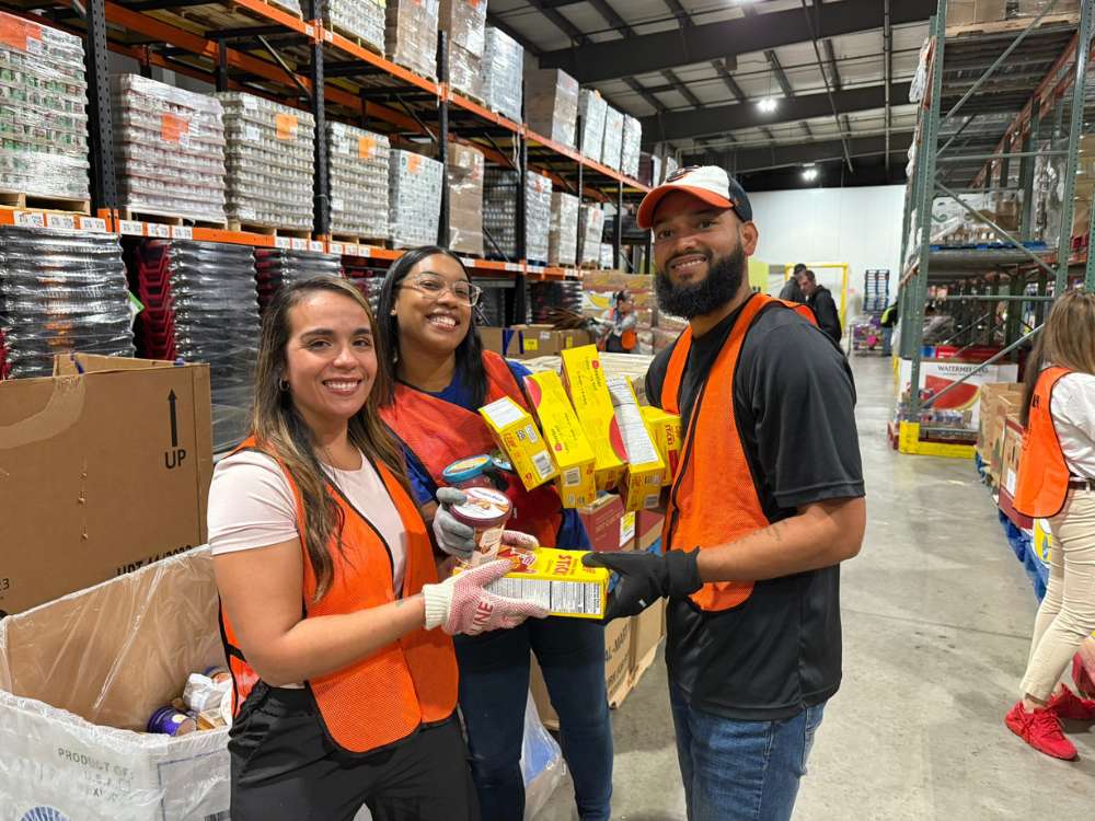 Three United Way of Berks County volunteers wearing bright orange safety vests smile while holding donated food items in a large warehouse. The group includes two women and a man, all holding various yellow food boxes and jars. In the background, tall industrial shelves are stacked high with pallets of canned goods and supplies.