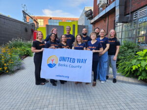 A group of ten people from United Way of Berks County standing in a paved outdoor courtyard. They are smiling and holding a large white banner featuring the United Way logo, the organization's name, and their website, uwberks.org. Many team members are wearing matching navy blue t-shirts that say "It starts with U" in orange script.