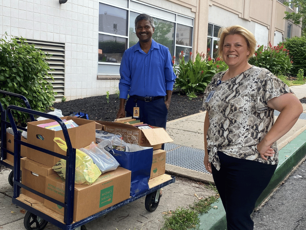 Gemini said Two United Way of Berks County volunteers, a man in a blue shirt and a woman in a snakeskin print top, stand smiling outdoors next to a blue handtruck loaded with several cardboard boxes and bags filled with items.