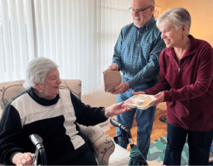 A seated elderly woman accepts a multi-compartment food tray from a smiling woman, as an older man stands holding a brown paper bag in a living room.
