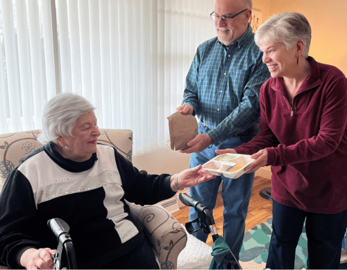 A seated elderly woman accepts a multi-compartment food tray from a smiling woman, as an older man stands holding a brown paper bag in a living room.