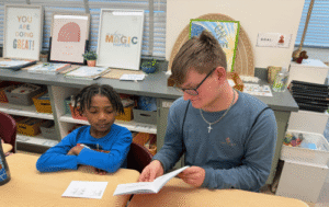 A young man in a blue shirt sits at a desk reading a book with a young student in a classroom.