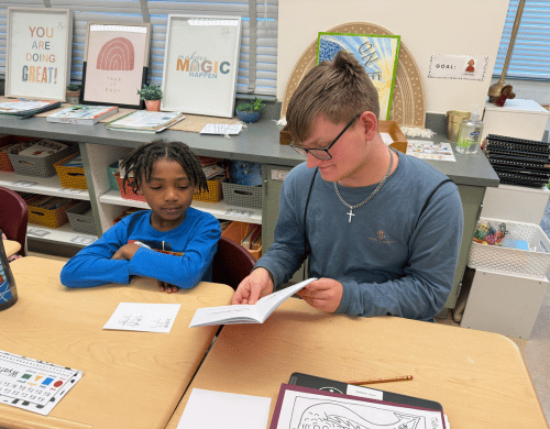 A young man in a blue shirt sits at a desk reading a book with a young student in a classroom.