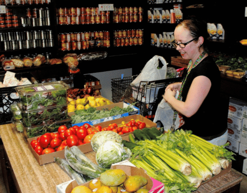 A woman selects fresh celery and tomatoes from a wooden table to pack into a grocery bag in a pantry stocked with canned goods.