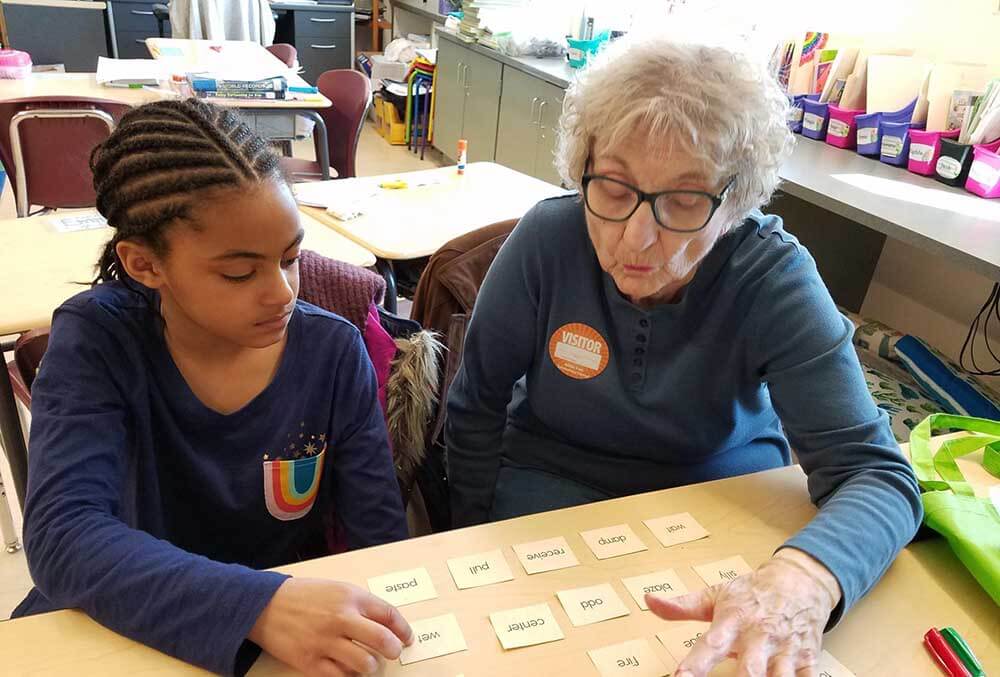 An older female volunteer wearing a visitor badge sits next to a young girl at a classroom desk. They are looking down and practicing reading using several small word cards spread across the table.