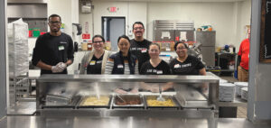 A group of men and women are standing behind a steam table in a commercial kitchen at a local shelter