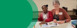 A second grade girl is smiling at the camera and making a peace sign with her fingers. Sitting next to her is a woman who is also smiling. They are both holding a book