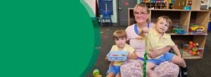 A grandmother is holding twin boys in her lap while sitting in a pre-K classroom. They are all smiling at the camera