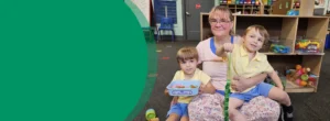 A grandmother is holding twin boys in her lap while sitting in a pre-K classroom. They are all smiling at the camera