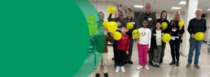 A group of adults and kids are standing in a school hallway. They are holding bright yellow balloons with words on them
