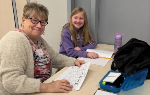A woman and a girl are sitting at a school table. They are holding worksheets and smiling