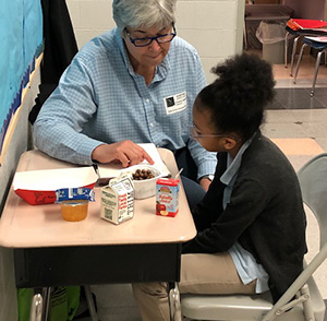 An adult volunteer reading tutor with gray hair and glasses, wearing a light blue button-down shirt, sits at a school desk next to a young girl with her hair in a high puff. The tutor is pointing to something in a book on the desk while providing reading tutoring as part of the Ready.Set.READ! program. In front of them on the desk are several school lunch items, including a carton of milk, a small juice box, a cup of fruit, and a container of food.