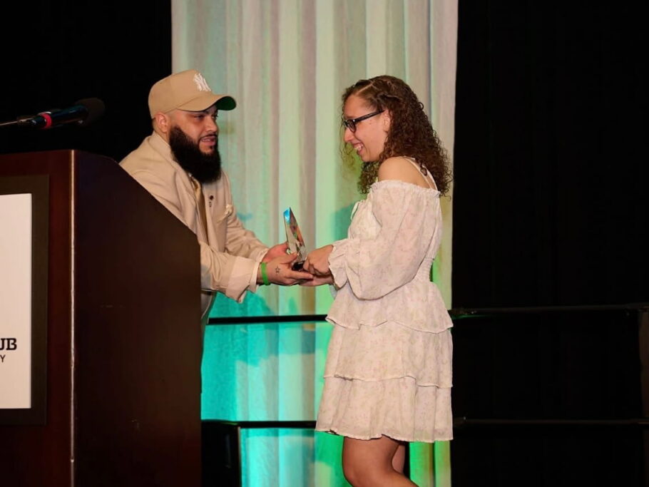 Zahavah smiling as she accepts a glass trophy from a presenter on stage next to the Olivet Boys & Girls Club podium.
