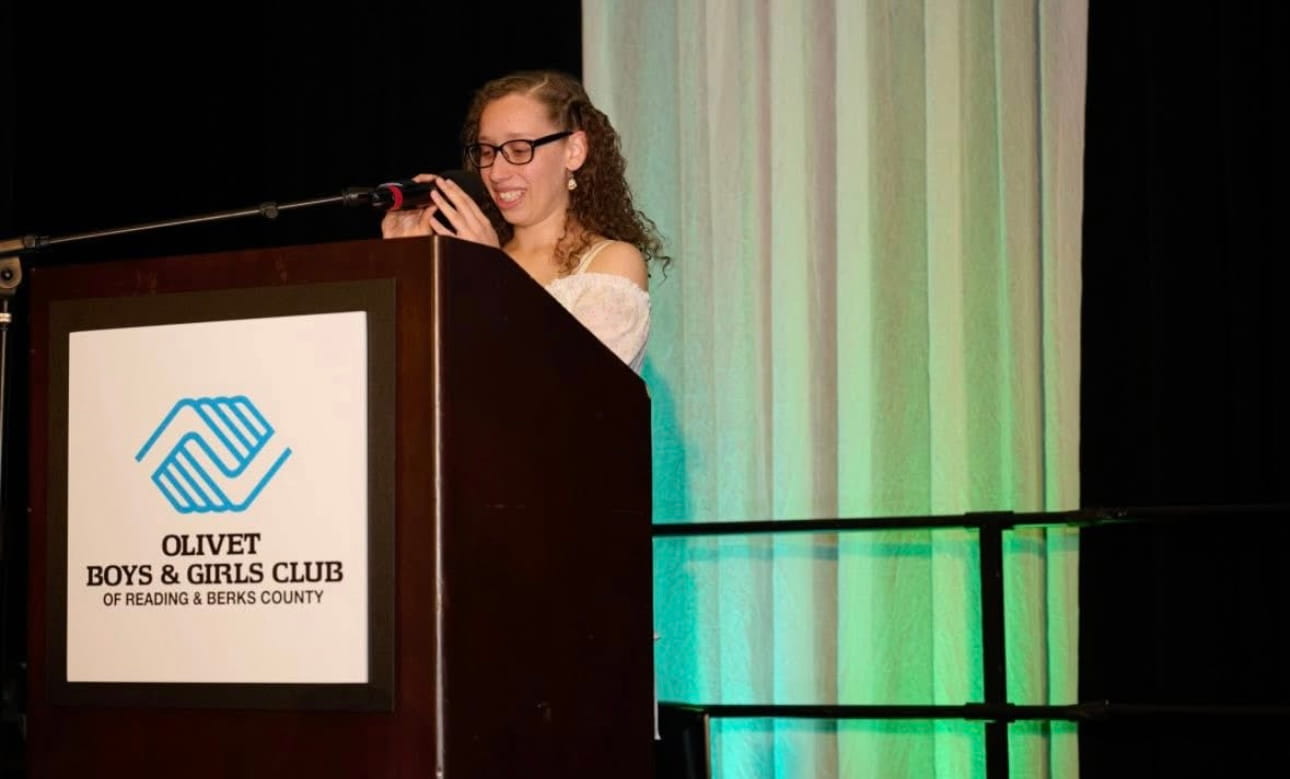 Zahavah smiling and speaking into a microphone at a wooden podium featuring the Olivet Boys & Girls Club logo.