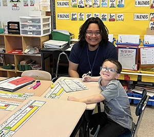 A teenage boy is sitting at a classroom table with an elementary-aged boy