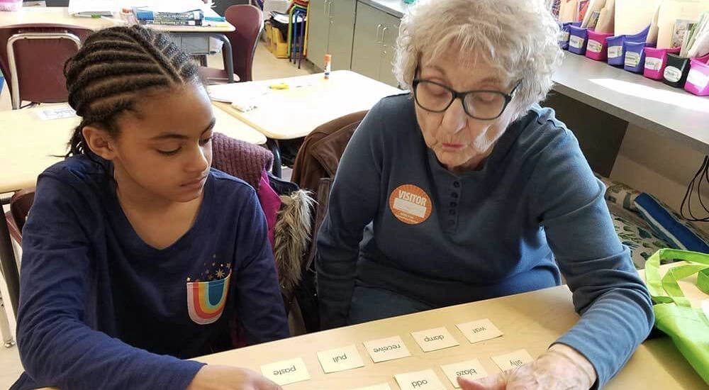 An older female volunteer wearing a visitor badge sits next to a young girl at a classroom desk. They are looking down and practicing reading using several small word cards spread across the table.